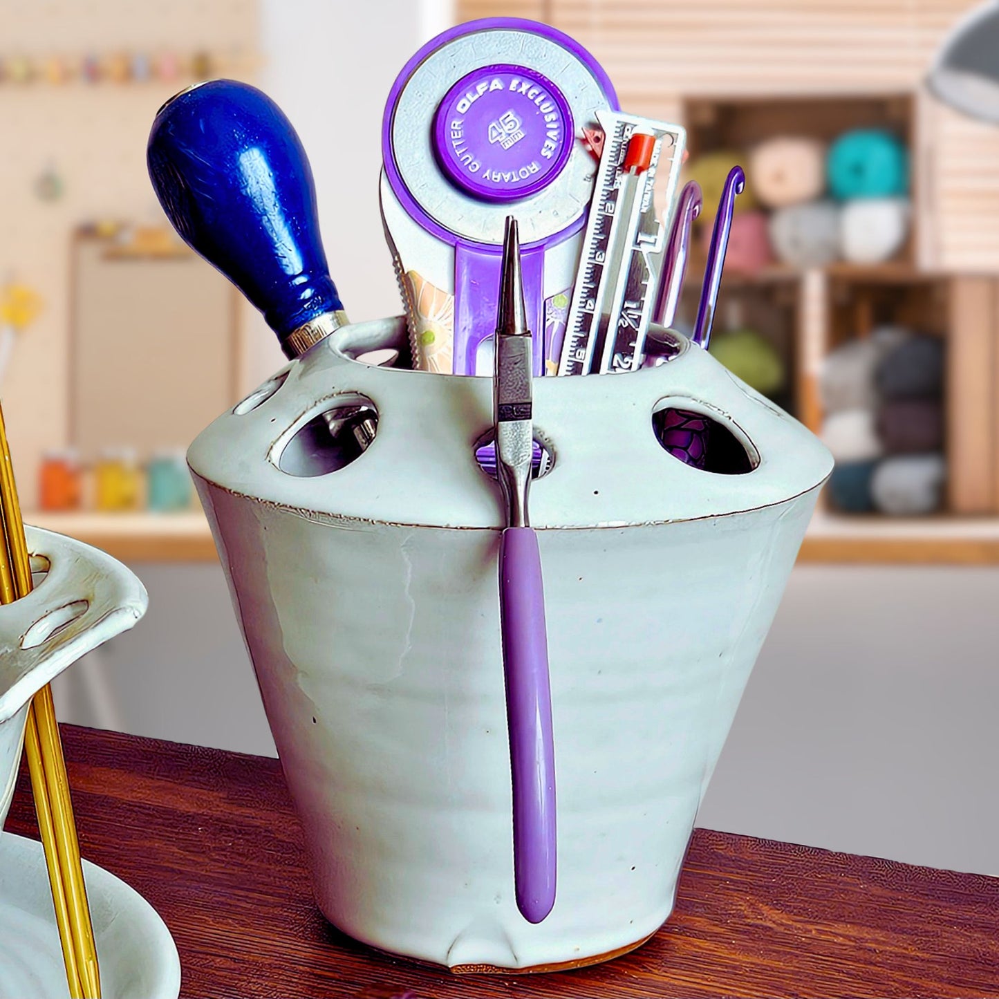 White ceramic organizer with various tools for crochet, knitting, fabric cutting, stitch measuring, crafting, jewelry making and leather work on a craft and sewing room table with pains and yarn in the background.