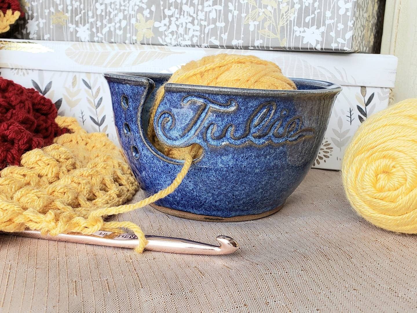 A custom ceramic yarn bowl with the name 'Julie' carved on the side, containing a skein of yellow yarn, alongside a partial view of a crochet piece and hook.