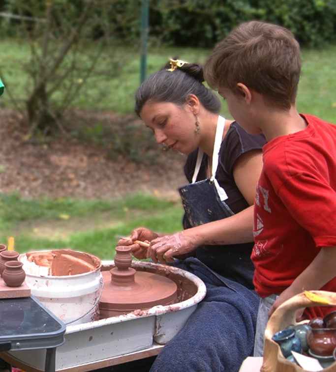Woman making tiny vases on the potter's wheel outside while a child watches