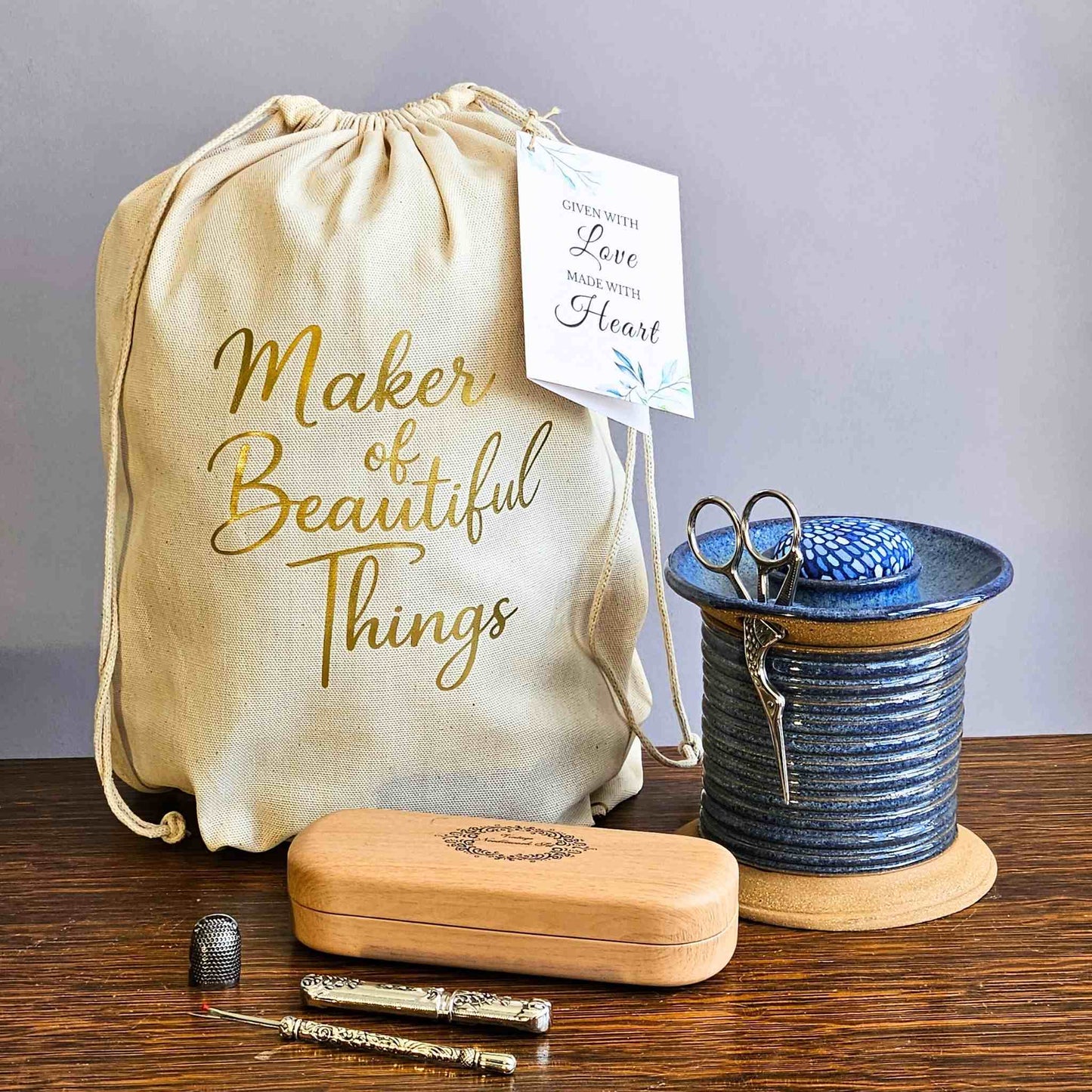 Sewing embroidery kit with a drawstring bag labeled 'Maker of Beautiful Things' on a wooden surface with assorted sewing notions, seam ripper, thimble, scissors, needle case, spool caddy.