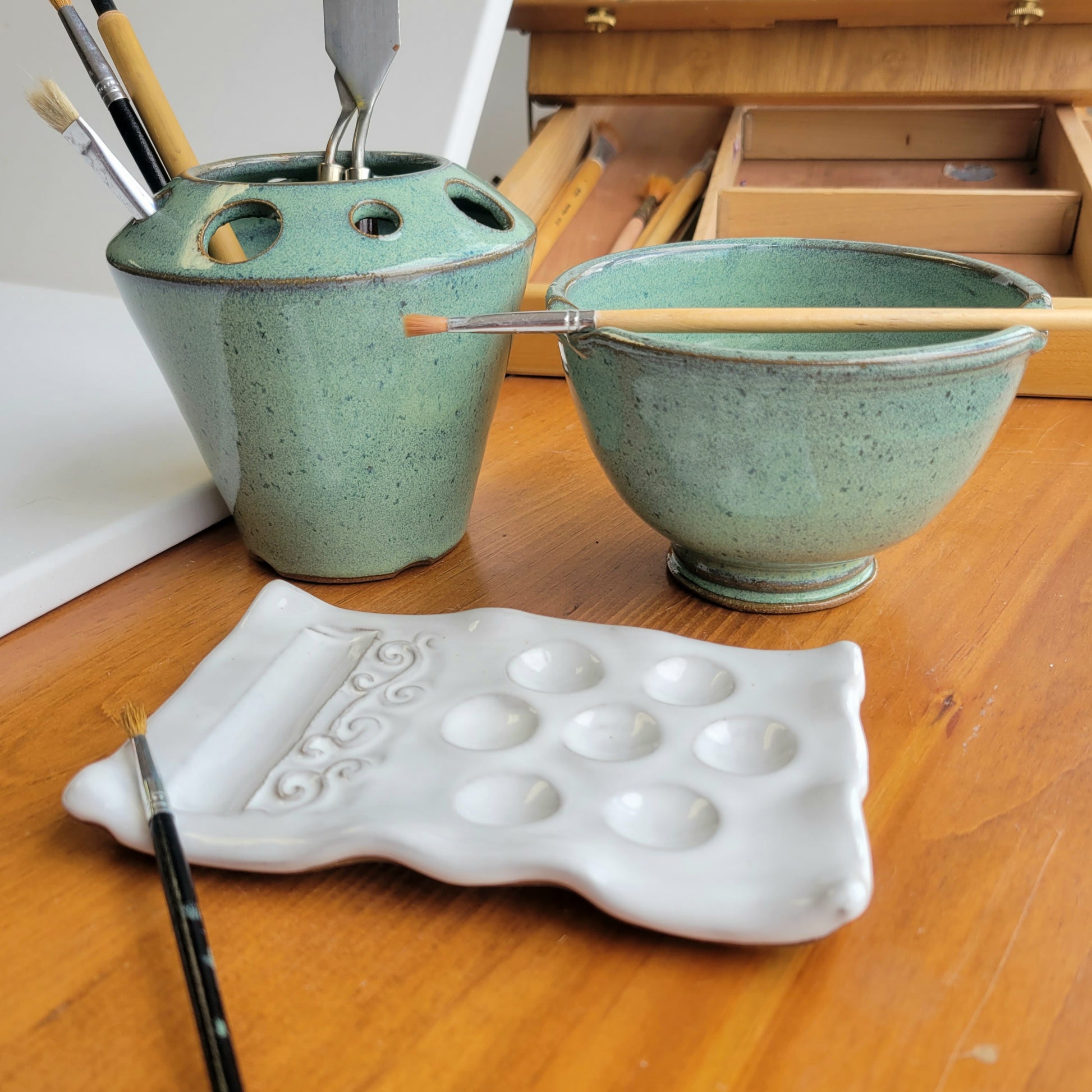 A handcrafted ceramic artist's gift set including a palette, water cup, and tool caddy in green, displayed on a wooden surface with brushes and a paintbrush resting on the tools.
