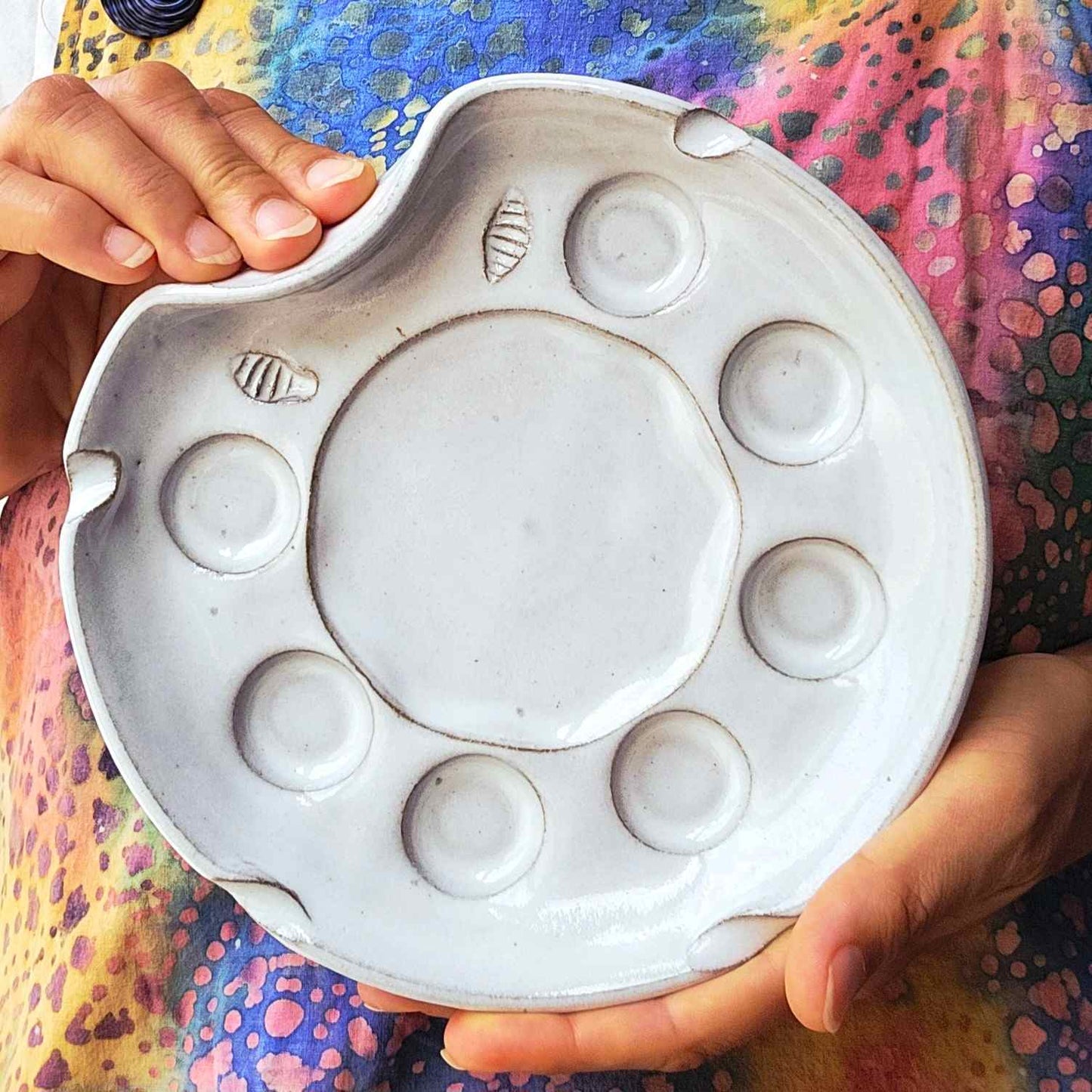 A ceramic paint palette with seven wells, large mixing area and brush rests, held by hands against a colorful artists apron. The palette has a white finish with brown speckles and small textured brush wiping area.