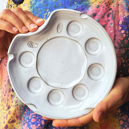 A ceramic paint palette with seven wells, large mixing area and brush rests, held by hands against a colorful artists apron. The palette has a white finish with brown speckles and small textured brush wiping area.