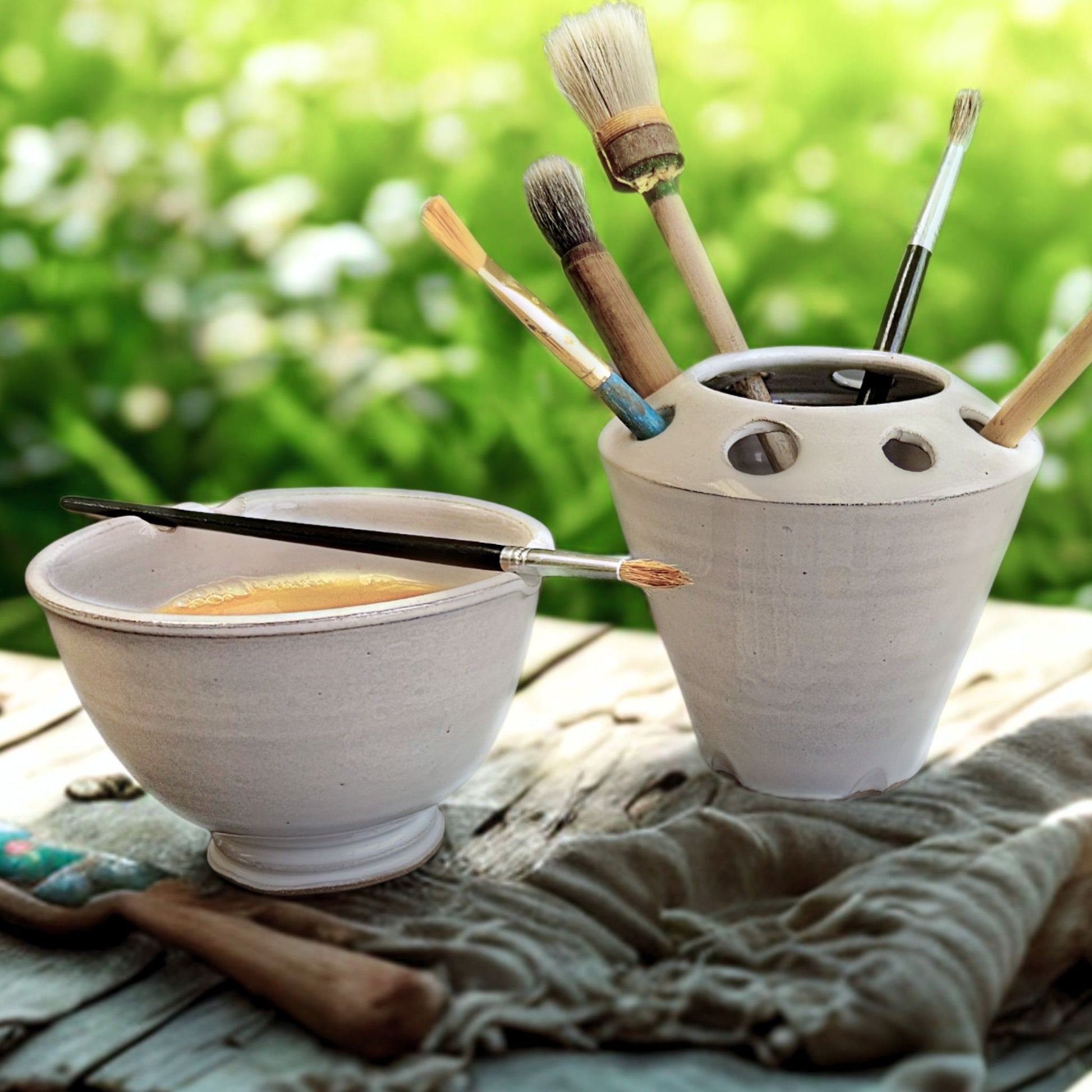 White ceramic painters water bowl and brush holder with paintbrushes on a wooden surface outdoors.