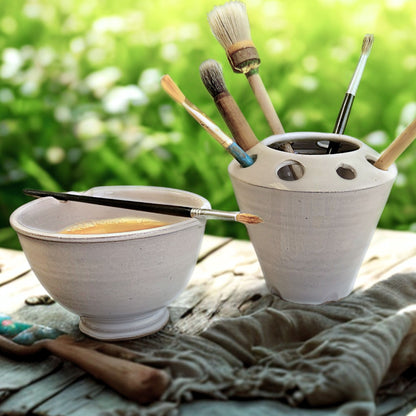White ceramic painters water bowl and brush holder with paintbrushes on a wooden surface outdoors.