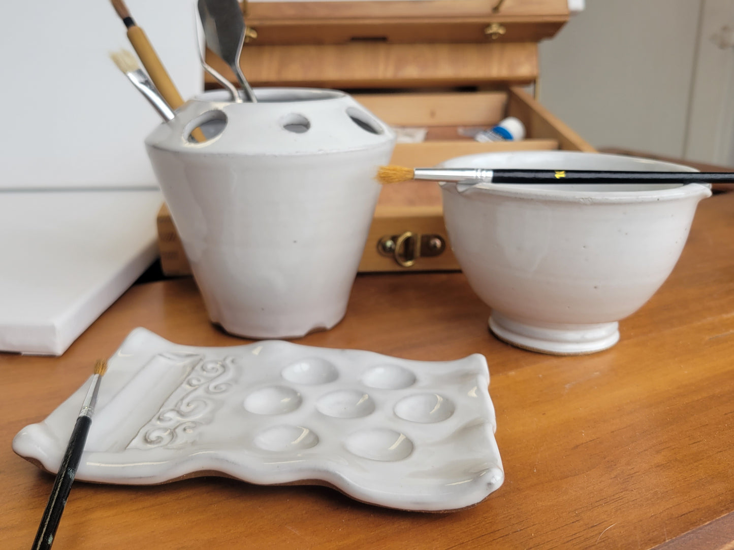 A handcrafted ceramic artist watercolor set including a paint palette, a brush caddy, and a brush rinse bowl, all in white color, displayed on a wooden surface.