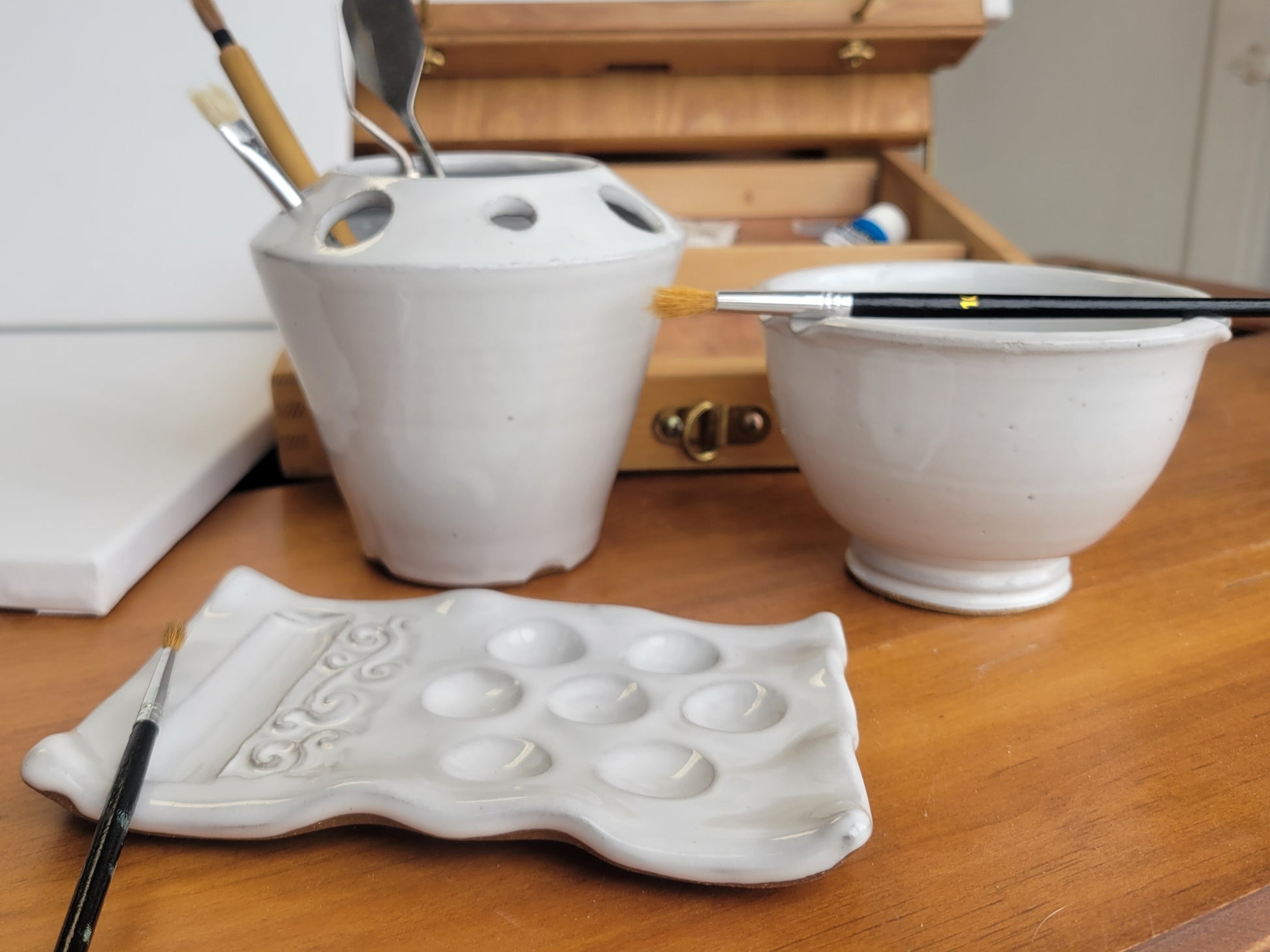 A handcrafted ceramic artist watercolor set including a paint palette, a brush caddy, and a brush rinse bowl, all in white color, displayed on a wooden surface.