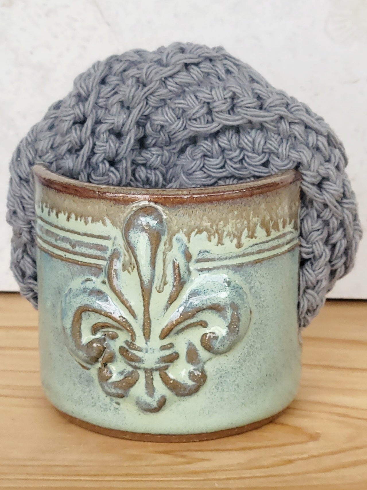 Front view of ceramic green sponge keeper with a decorative fleur-de-lis design holding a crochet cotton yarn scrubber sponge. Perfect for kitchen counter organization. 