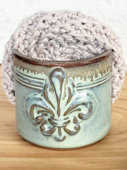 Front view of ceramic green sponge keeper with a decorative fleur-de-lis design holding a crochet cotton yarn scrubber sponge. Perfect for kitchen counter organization. 