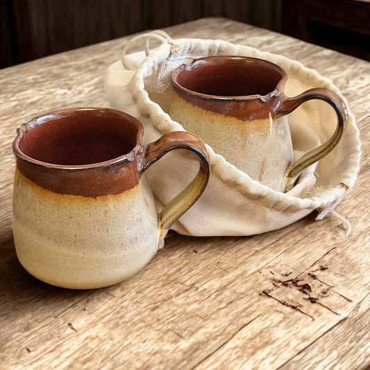 Two ceramic mugs with brown accents in a canvase drawstring coffee bean sack on a wooden surface.