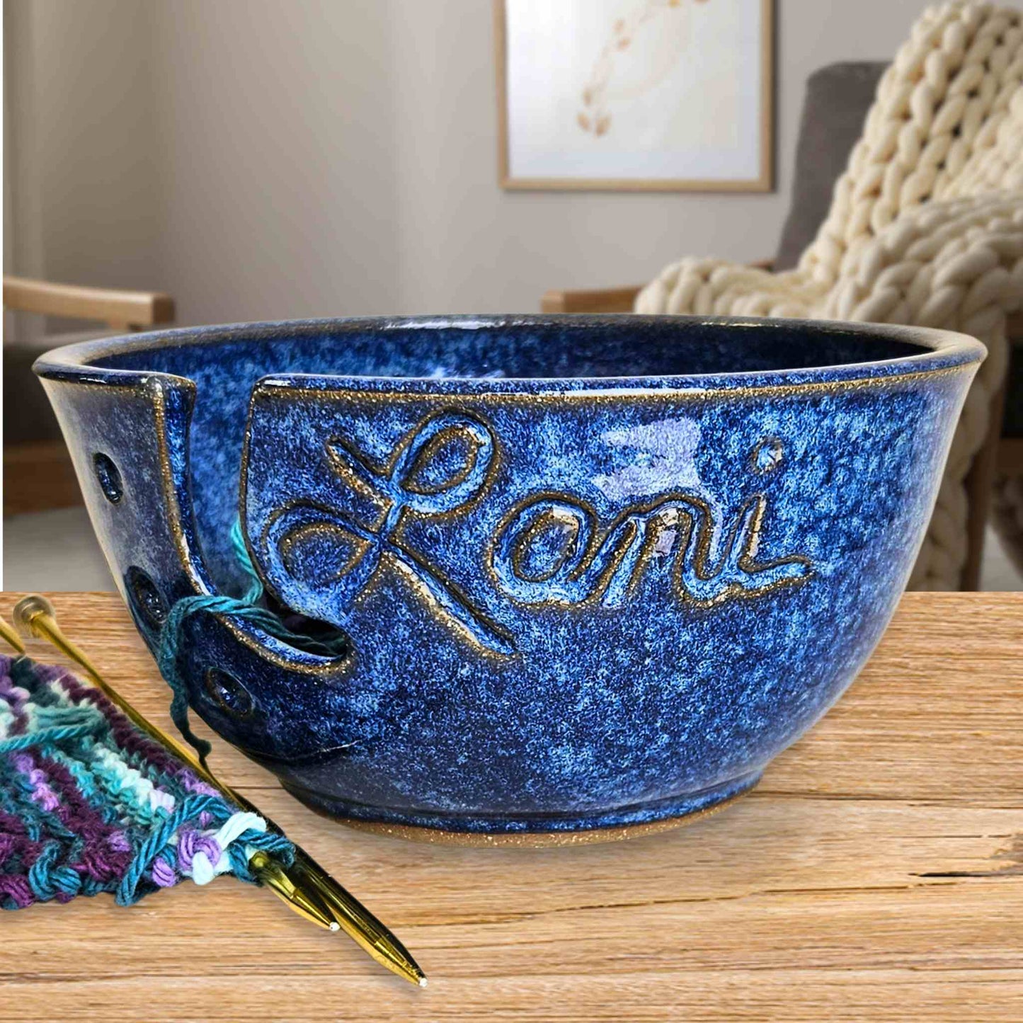 Custom name blue ceramic yarn bowl holding a knitting project with holes and the name Lani carved into the front on a wooden table and knitted blanket in the background
