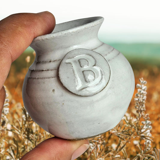 white tiny handmade pottery vase with letter B embossed into it held between fingers to showcase its miniature size with a background of a field of fall wild flowers with the ocean in the far distance.