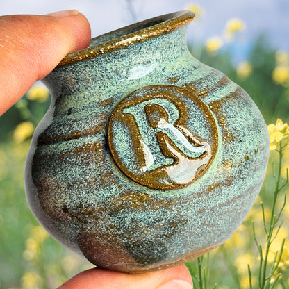 Green miniature handmade pottery vase with letter R engraved into it held between thumb and forefinger to showcase its small size with a background of a field of yellow wildflowers.