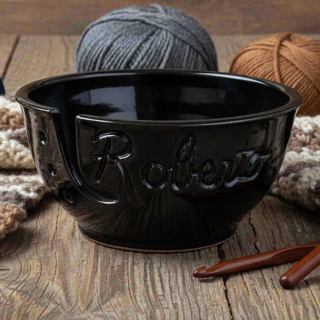ceramic yarn bowl in black with name 'Roberto' carved into it on a wooden table with crochet hooks and skeins of brown and grey yarn.