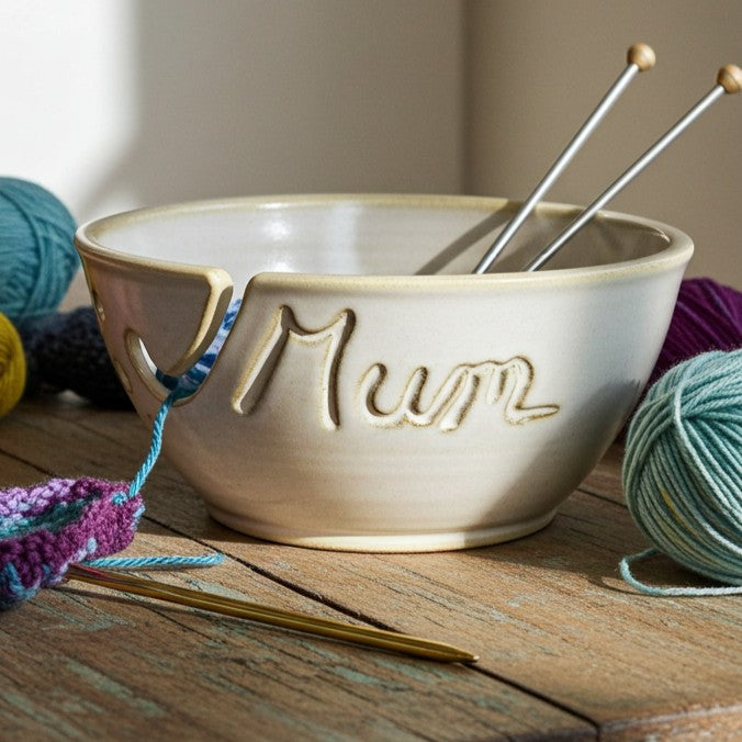 Custom yellow ceramic yarn bowl with hand-carved name 'Mum' on table with yarn and knitting needles. Handmade pottery knitting bowl with yarn guide slot and needle holes. Personalized crochet gift for Mother's Day, Christmas for any mom, grandmother, aunt or friend. 