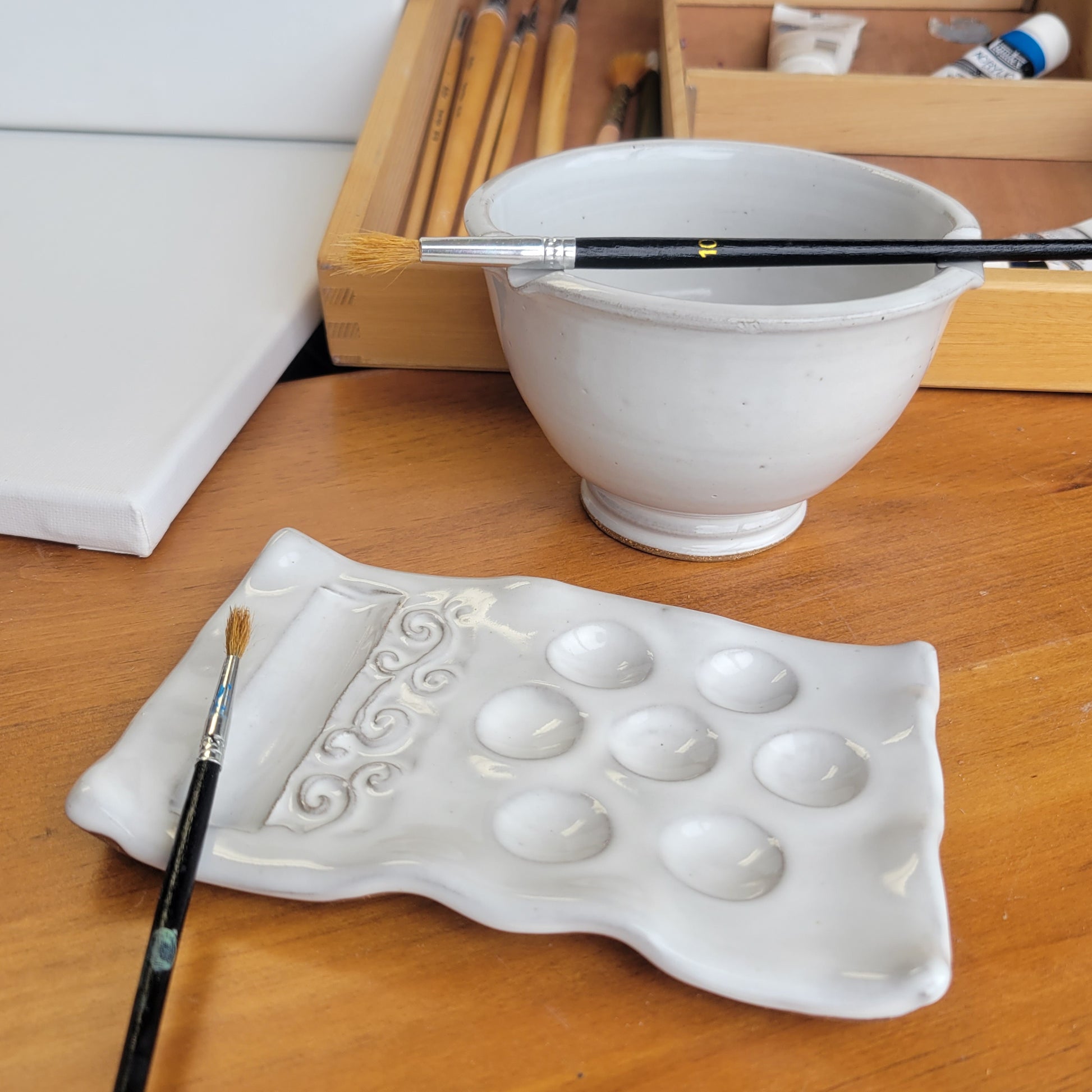 A handcrafted ceramic artist watercolor set including a paint palette, a brush caddy, and a brush rinse bowl, all in white color, displayed on a wooden surface.