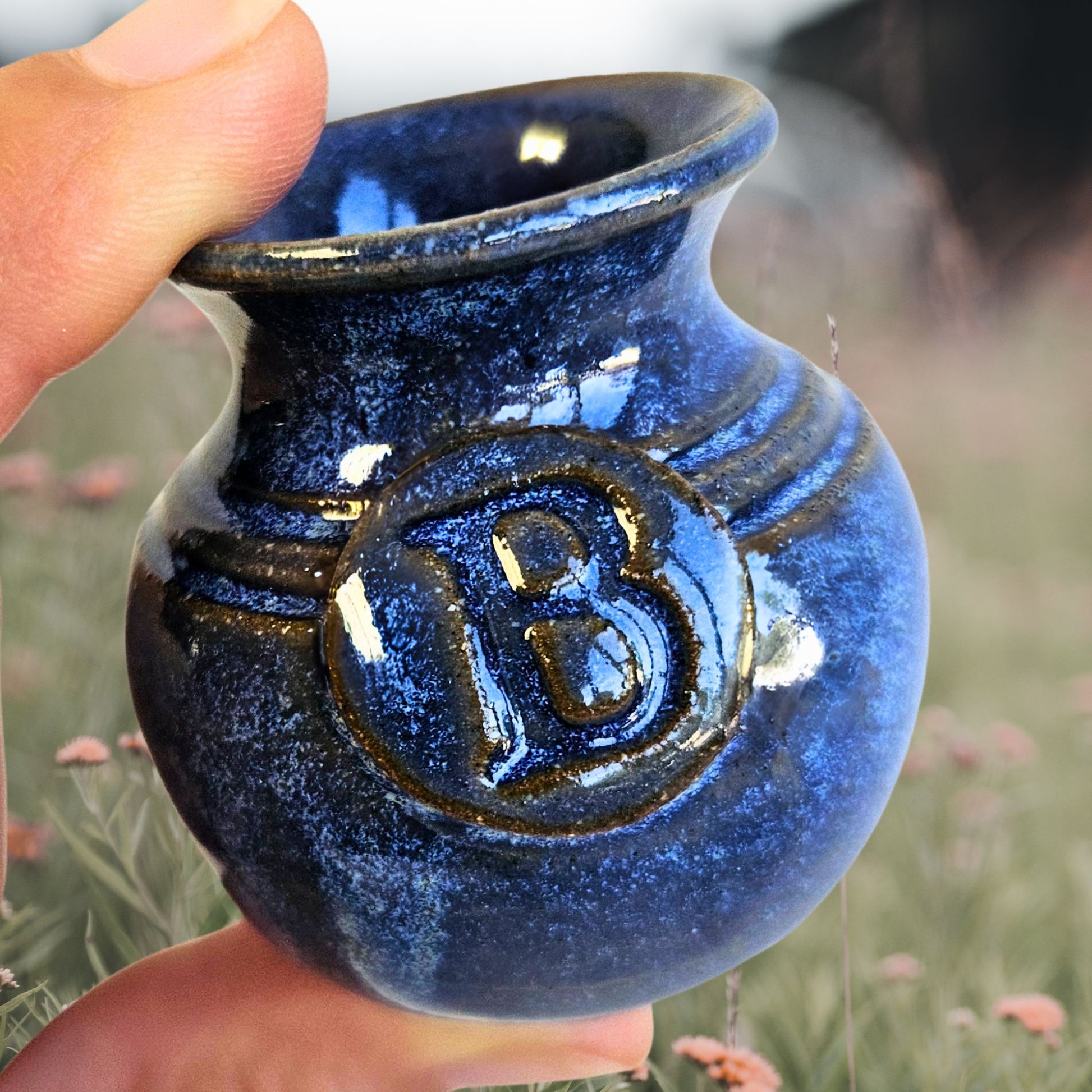 blue personalized handmade pottery vase with letter B engraved into it, held between fingers to showcase its miniature size with a background of a field of wild flowers.