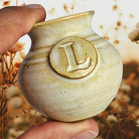 Cream colored small handmade pottery vase with letter L stamped into it on a plaque and held between fingers to showcase its miniature size with a background of a field of fall flowers at sunset.