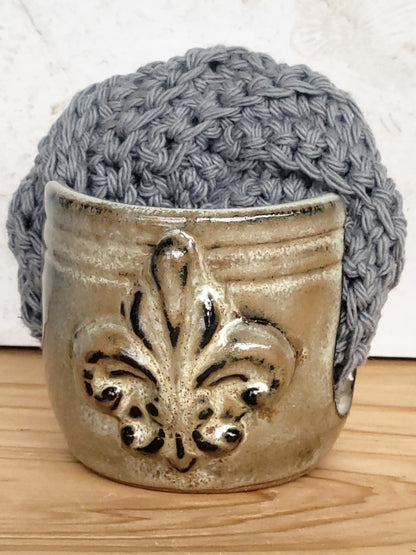 Handmade artisan bronze ceramic sponge holder with embossed fleur-de-lis design, paired with a dark gray cotton crochet scrubber sponge. Eco-friendly kitchen countertop accessory perfect for storing crochet scrubbers, dish sponges, and sink cleaning essentials.