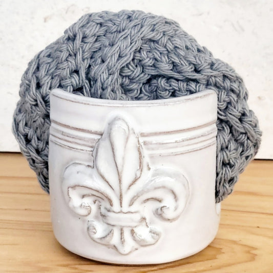 Ceramic white sponge keeper with a decorative fleur-de-lis design holding a crochet cotton yarn scrubber sponge. Perfect for kitchen counter organization. 
