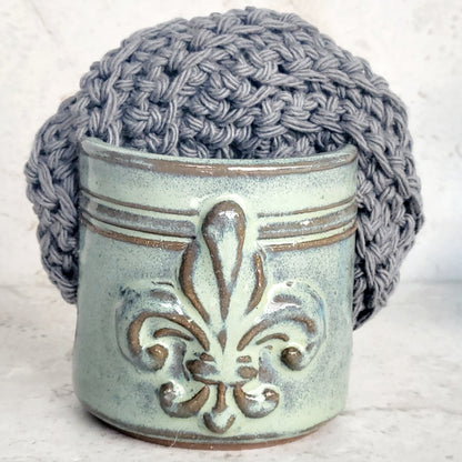 Ceramic sponge keeper with a decorative fleur-de-lis design holding a crochet cotton yarn scrubber sponge.