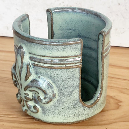 Side view of a green ceramic sponge holder with decorative fleur de lis pattern on a wooden surface
