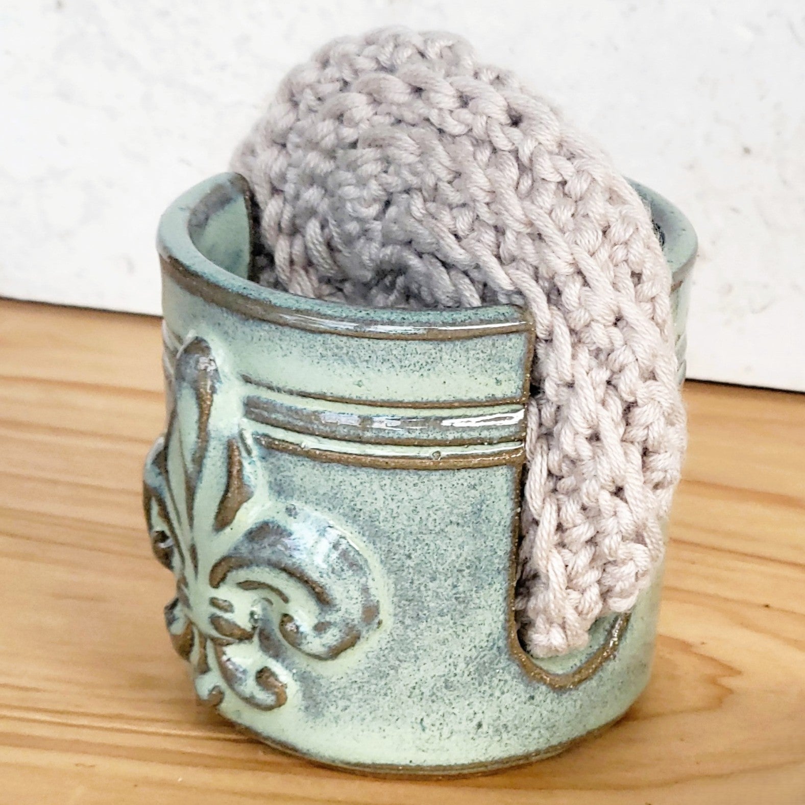 side view of a decorative fleur de lis embossed sponge holder with a beige crochet scrubber in it on a wooden countertop