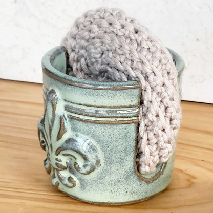 side view of a decorative fleur de lis embossed sponge holder with a beige crochet scrubber in it on a wooden countertop