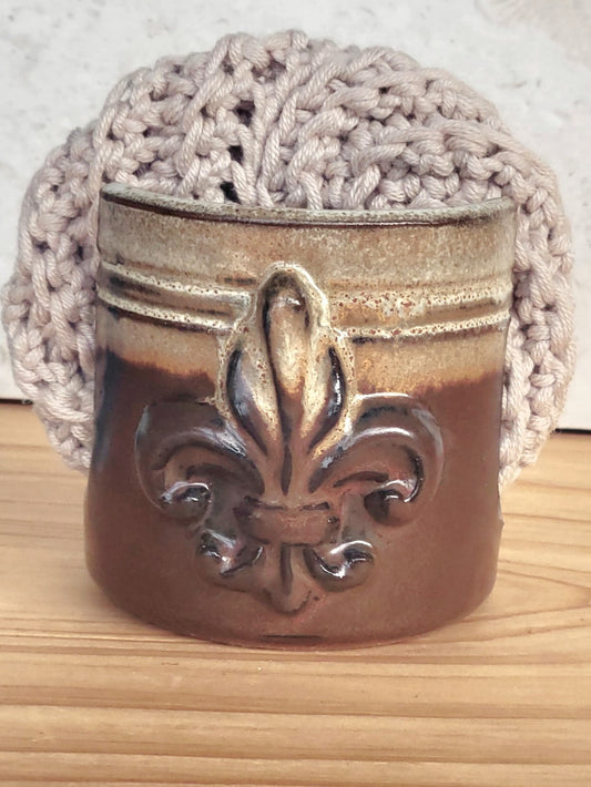 Front view of ceramic brown sponge keeper with a decorative fleur-de-lis design holding a crochet cotton yarn scrubber sponge. Eco-friendly kitchen countertop accessory.