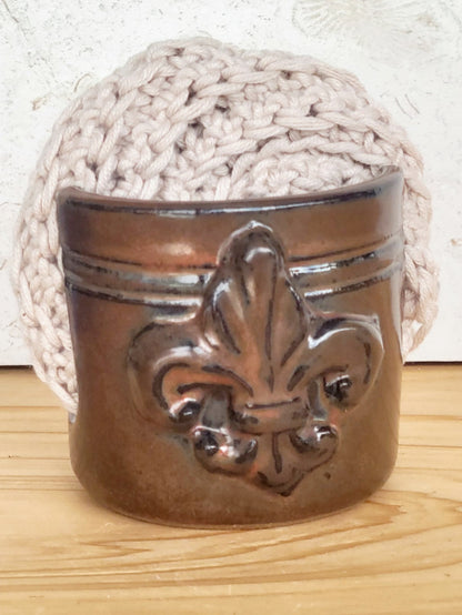Brown handmade ceramic sponge keeper with a decorative fleur-de-lis design holding a crochet cotton yarn scrubber sponge. Stylish kitchen sink organizer and eco-friendly countertop accessory.