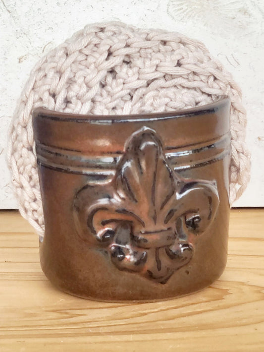 Brown handmade ceramic sponge keeper with a decorative fleur-de-lis design holding a crochet cotton yarn scrubber sponge. Stylish kitchen sink organizer and eco-friendly countertop accessory.