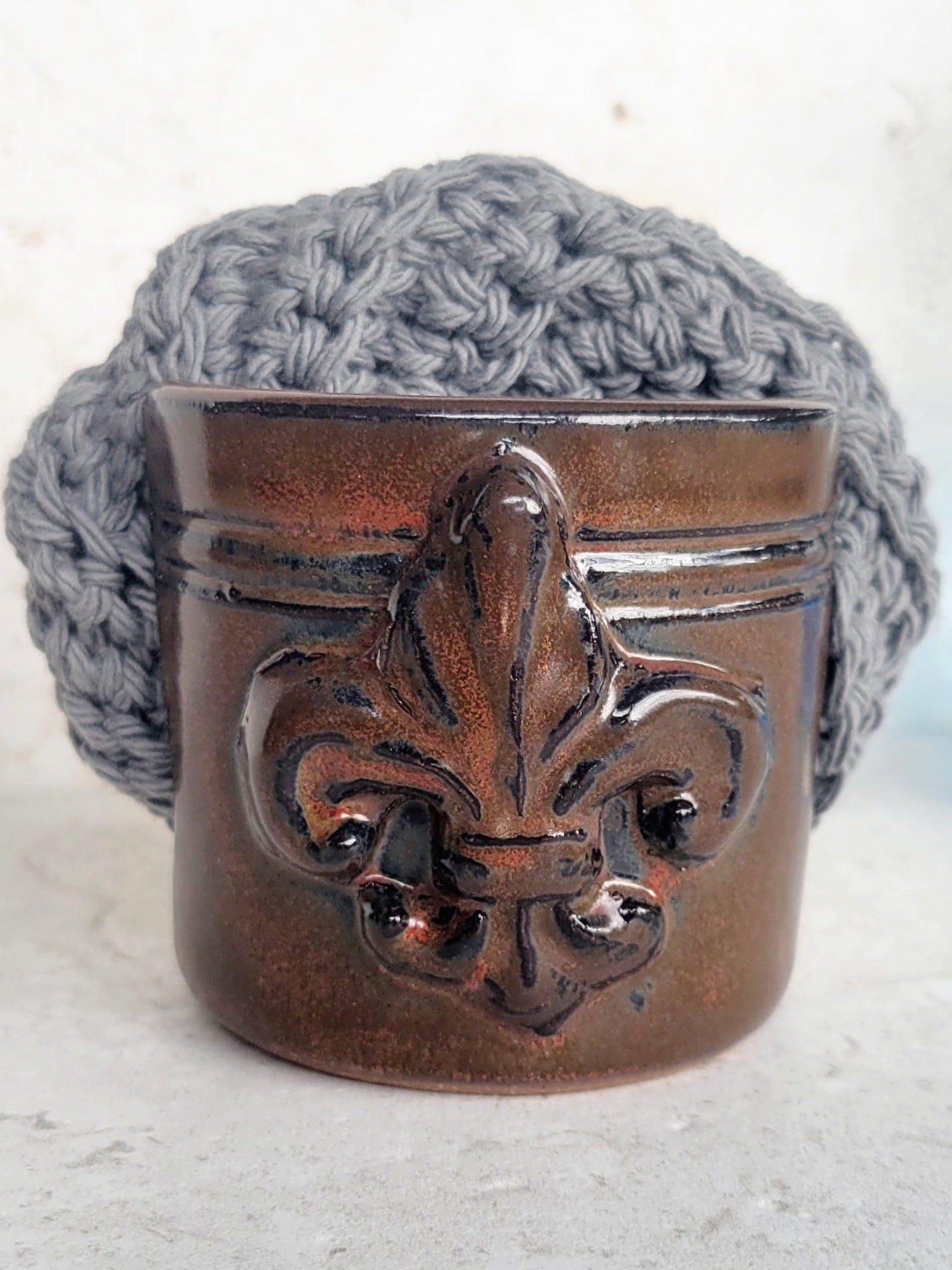 Brown handmade ceramic sponge keeper with a decorative fleur-de-lis design holding a gray crochet cotton yarn scrubber sponge. Stylish kitchen sink organizer and eco-friendly countertop accessory.