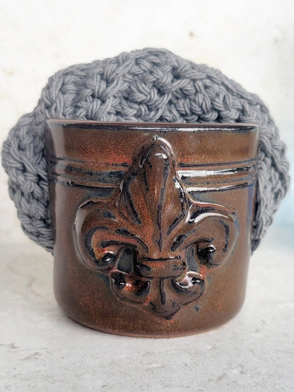 Brown handmade ceramic sponge keeper with a decorative fleur-de-lis design holding a gray crochet cotton yarn scrubber sponge. Stylish kitchen sink organizer and eco-friendly countertop accessory.