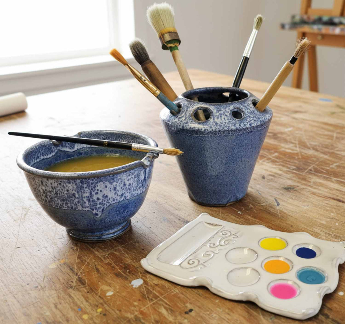 A paintbrush holder and matching paint brush rinse bowl on a wooden table, alongside a white paint palette with colorful wells and several paintbrushes and made of ceramic. 