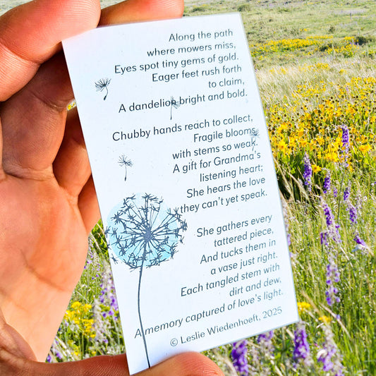 Hand holding a printed card with a poem and dandelion illustration against a background of a field of wild flowers.