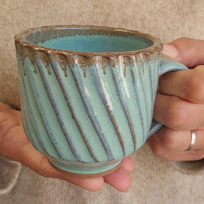 Hand holding a ceramic mug with a diagonally striped textured surface and green bronze glaze.