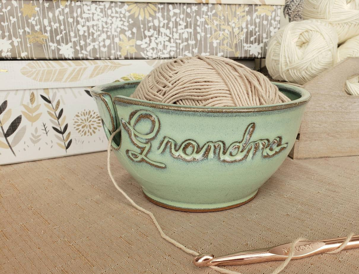 A custom ceramic yarn bowl with the word 'Grandma' carved on the side, containing a skein of beige yarn, alongside a partial view of a knitted piece.