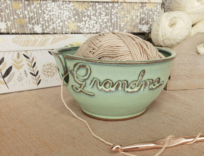 A custom ceramic yarn bowl with the word 'Grandma' carved on the side, containing a skein of beige yarn, alongside a partial view of a knitted piece.