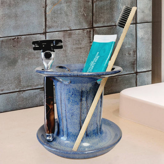 A blue handmade ceramic toothbrush stand with multiple slots, displaying a toothbrush, toothpaste tube, and a razor. The stand has a striped pattern and is placed on a light-colored bathroom counter. Multi-purpose bathroom organizer keeps oral care essentials neat and accessible.