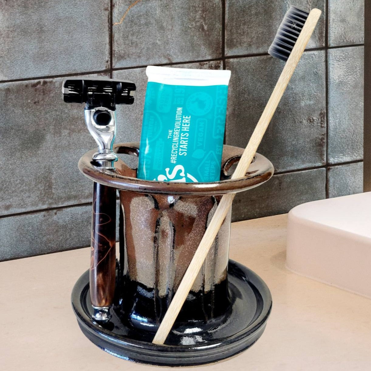 A black and brown handmade pottery toothbrush stand with multiple slots, displaying a toothbrush, toothpaste tube, and a razor. The stand has a striped pattern and is placed on a light-colored bathroom counter. Functional bathroom organizer for toothpaste, shaving gear, and grooming essentials.