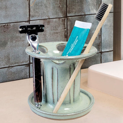 A green handmade ceramic toothbrush stand with multiple slots, displaying a toothbrush, toothpaste tube, and a razor. The stand has a striped pattern and is placed on a light-colored bathroom counter. Functional bathroom organizer for toothpaste, shaving gear, and essentials.
