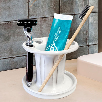 A white handmade ceramic toothbrush stand with multiple slots, displaying a toothbrush, toothpaste tube, and a razor. The stand has a striped pattern and is placed on a light-colored bathroom counter. Functional bathroom organizer for toothpaste, shaving gear, and grooming essentials.
