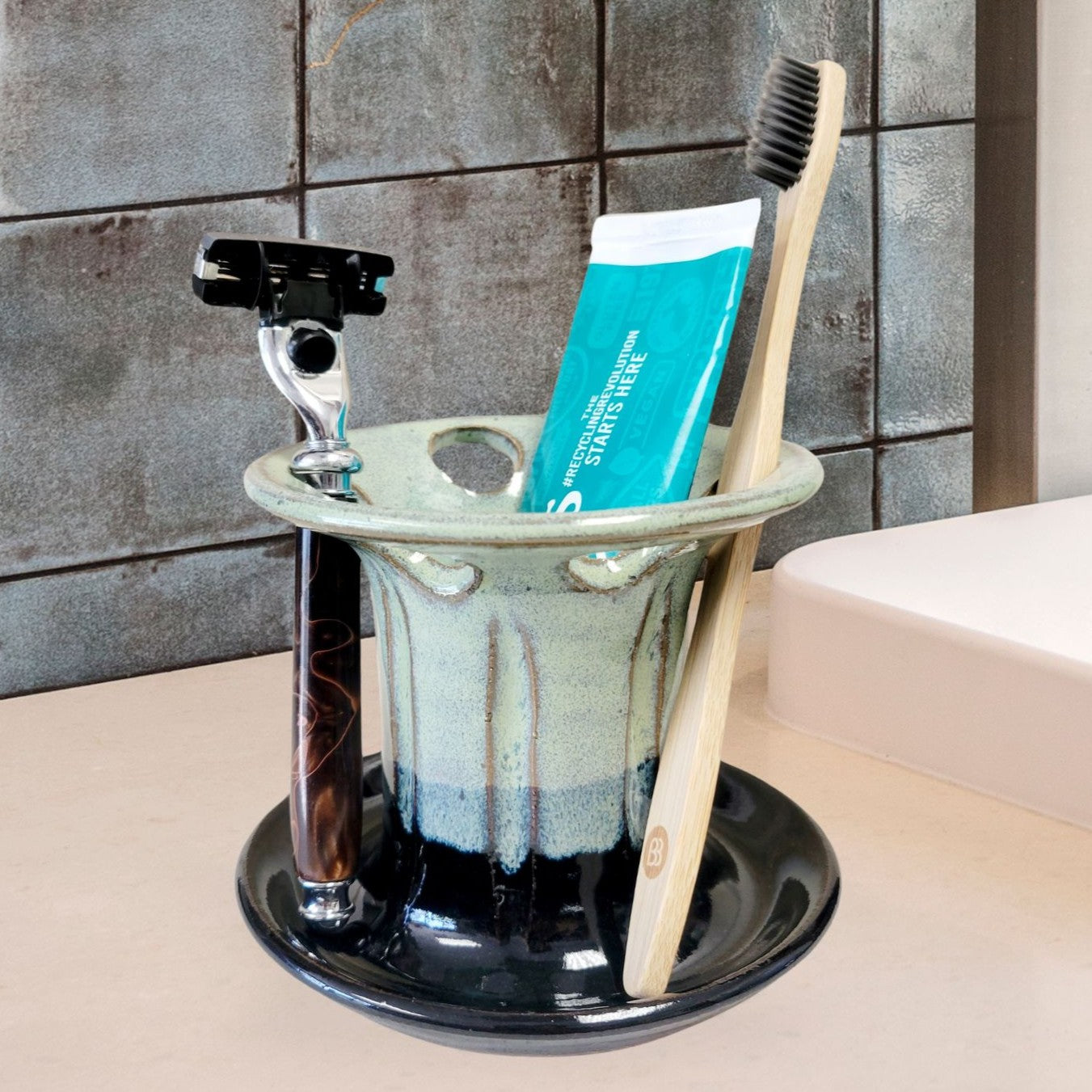 A green and black handmade ceramic toothbrush stand with multiple slots, displaying a toothbrush, toothpaste tube, and a razor. The stand has a striped pattern and is placed on a light-colored bathroom counter. Handmade ceramic bathroom organizer for countertop storage.