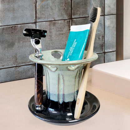 A green and black handmade ceramic toothbrush stand with multiple slots, displaying a toothbrush, toothpaste tube, and a razor. The stand has a striped pattern and is placed on a light-colored bathroom counter. Handmade ceramic bathroom organizer for countertop storage.