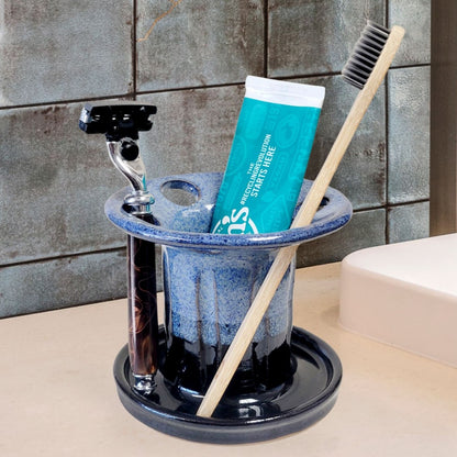 A blue and black handmade ceramic toothbrush stand with multiple slots, displaying a toothbrush, toothpaste tube, and a razor. The stand has a striped pattern and is placed on a light-colored bathroom counter. Handmade ceramic bathroom organizer for countertop storage.