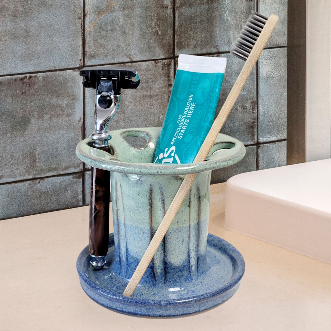 A handmade teal and blue ceramic toothbrush stand with multiple slots, displaying a toothbrush, toothpaste tube, and a razor. The stand has a striped pattern and is placed on a light-colored bathroom counter. Stylish bathroom countertop organizer.