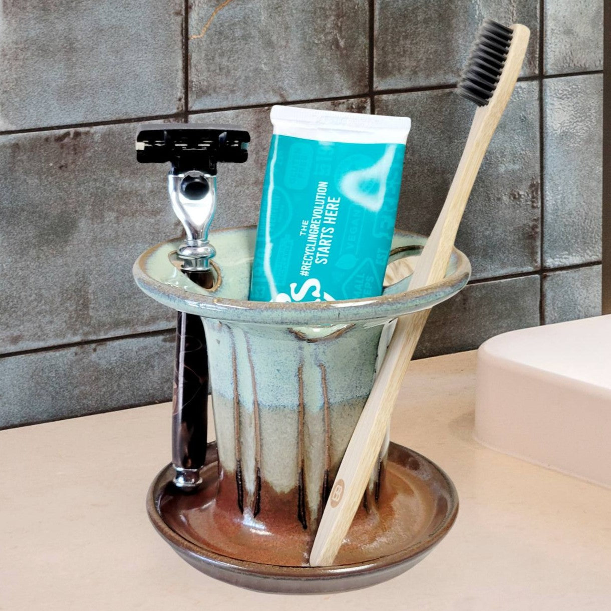 A green and brown handmade ceramic toothbrush stand with multiple slots, displaying a toothbrush, toothpaste tube, and a razor. The stand has a striped pattern and is placed on a light-colored bathroom counter. Handmade ceramic bathroom organizer for countertop storage.