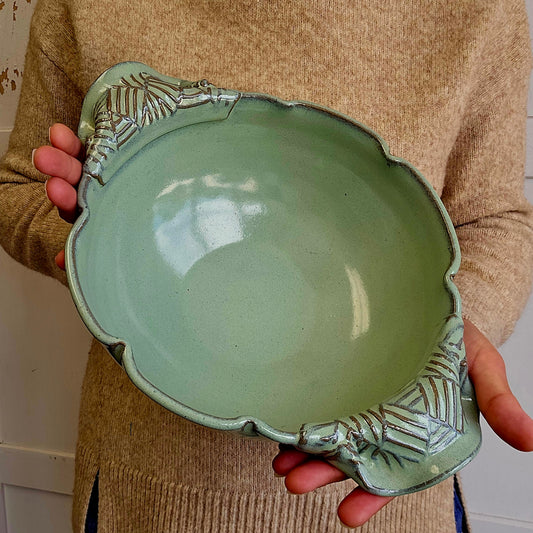 Person holding a green ceramic bowl with decorative handles and a scalloped rim.