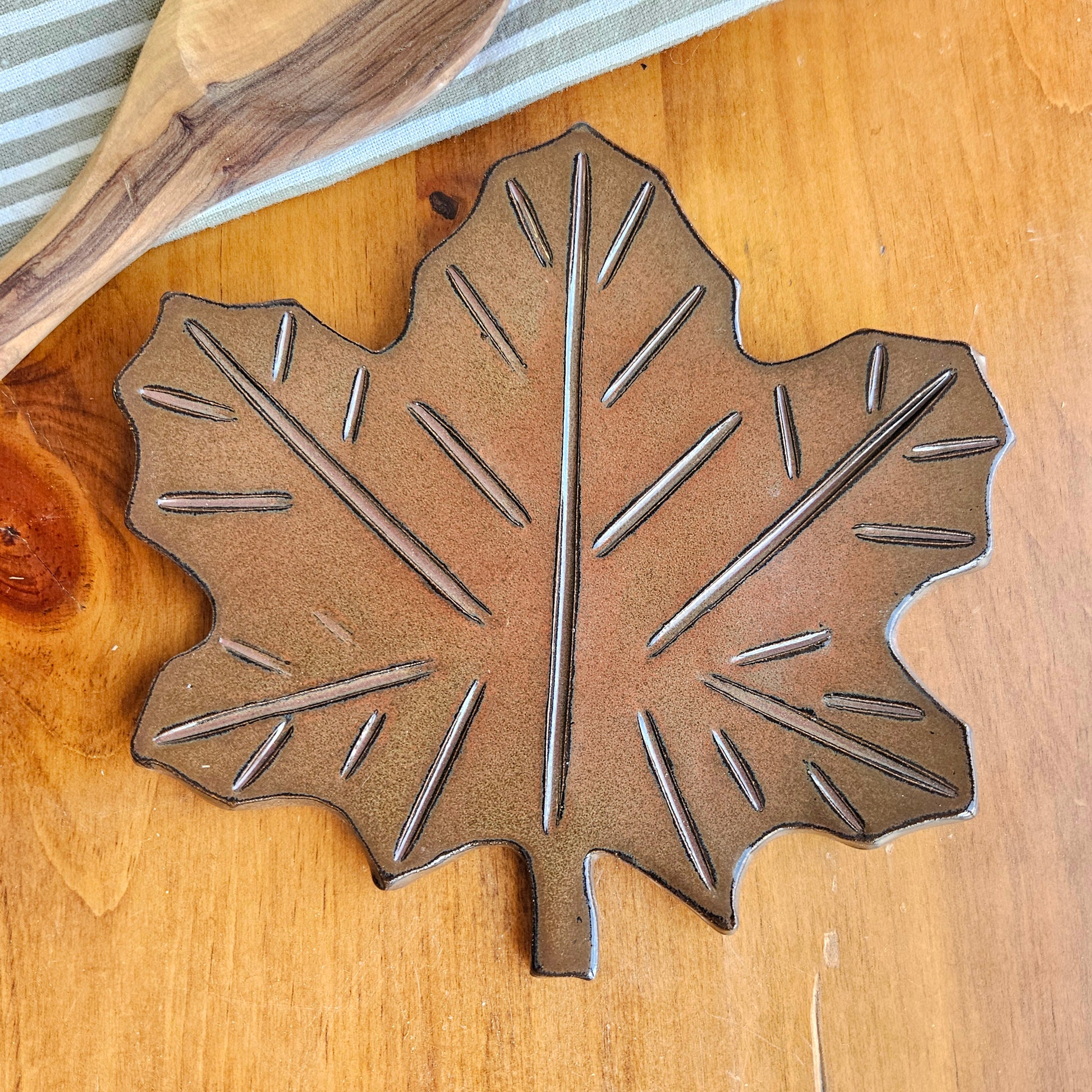 Handmade rust-colored maple leaf ceramic spoon rest on rustic kitchen counter – fall-inspired pottery decor piece next to a wooden mixing spoon and cotton kitchen towel.