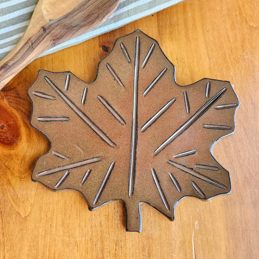 Handmade rust-colored maple leaf ceramic spoon rest on rustic kitchen counter – fall-inspired pottery decor piece next to a wooden mixing spoon and cotton kitchen towel.